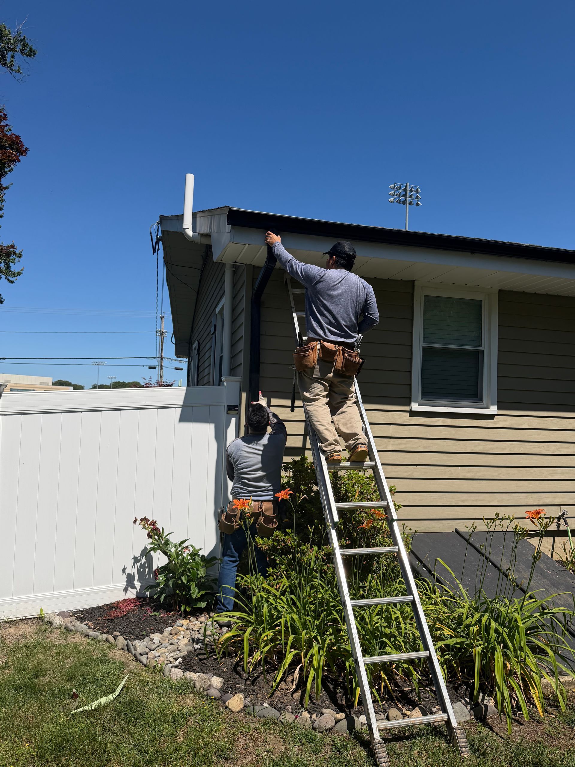 Two workers on a ladder repairing the roof edge of a house under a clear blue sky