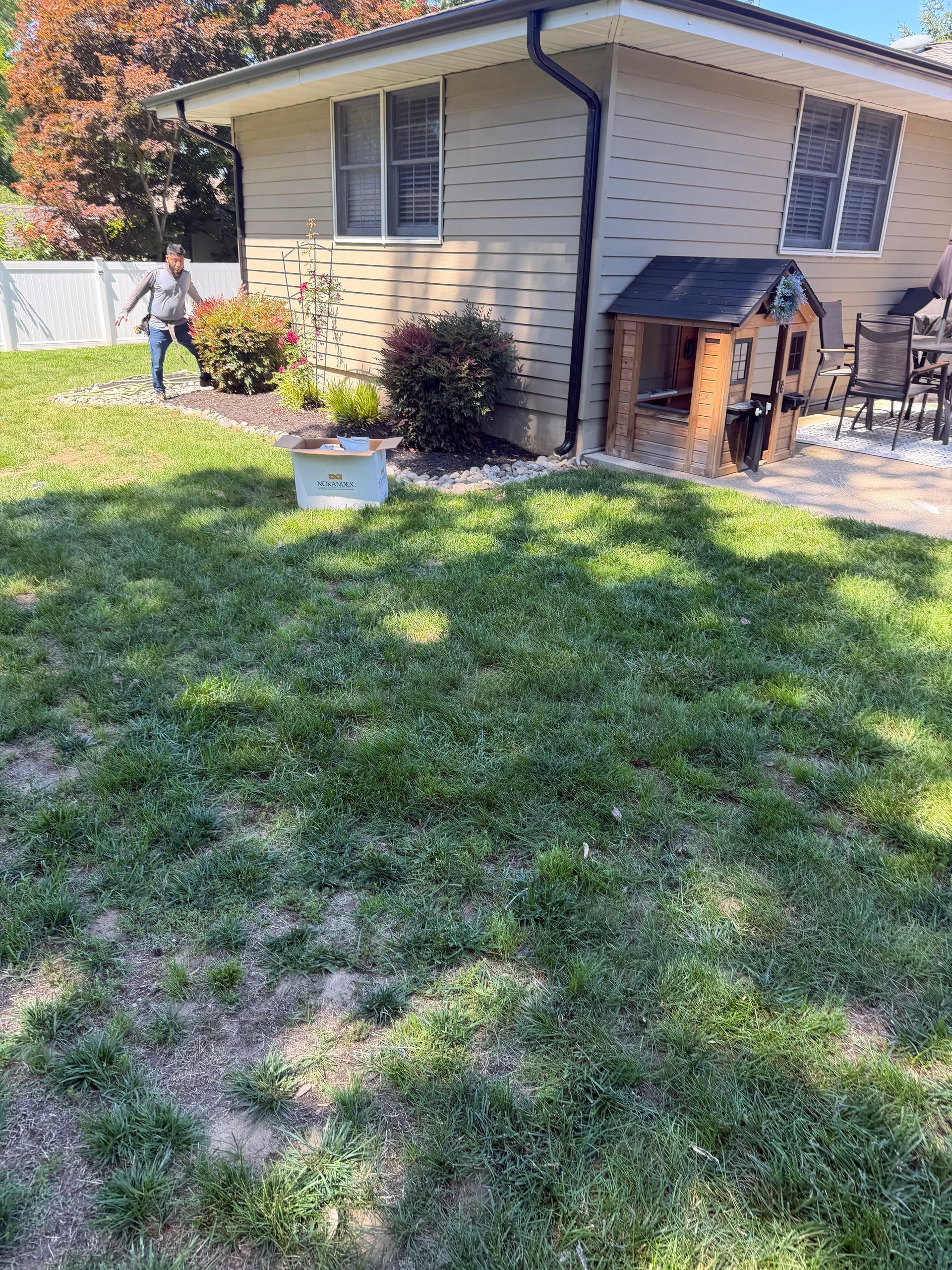 Backyard with a beige house, green lawn, and a person near a white fence in the background