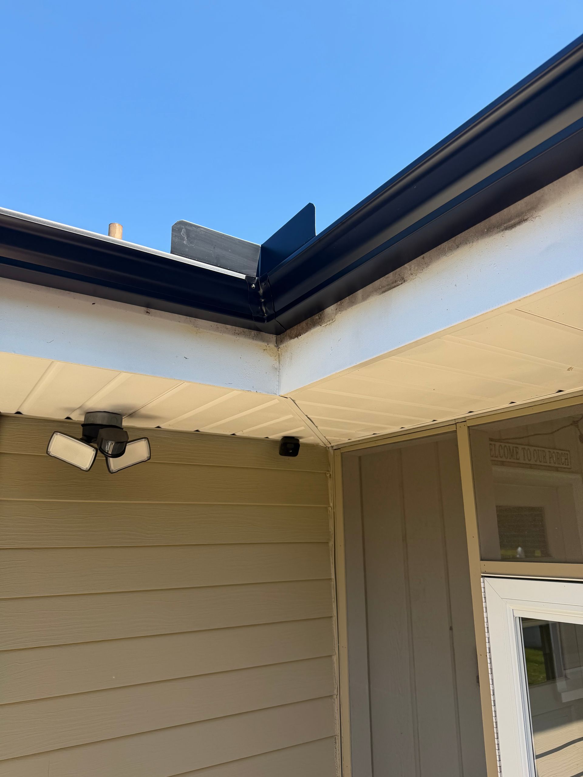Exterior corner of a house with beige siding, white trim, a security light, and a blue sky overhead