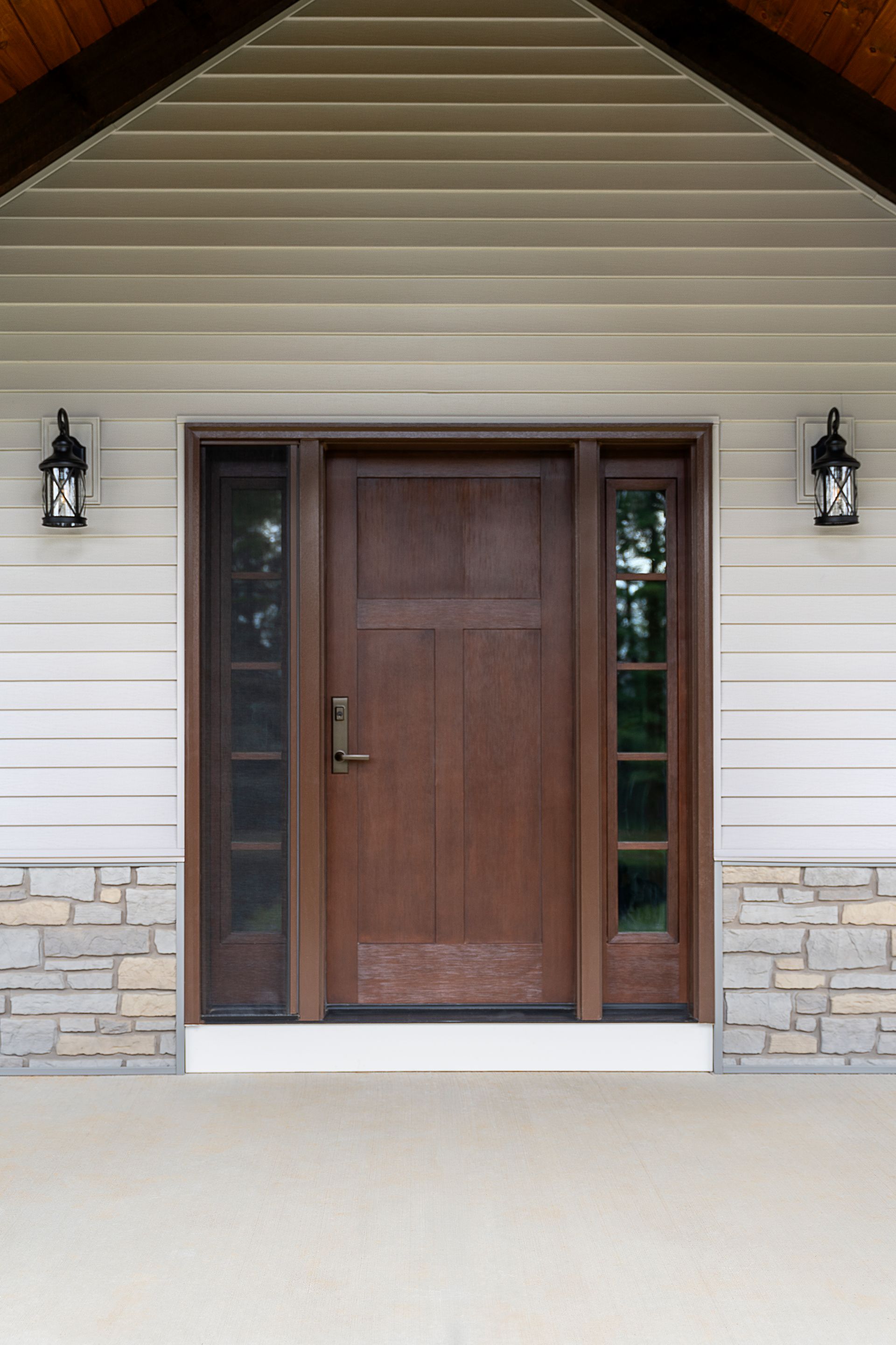 Front porch entry with a wooden door, sidelights, and wall lanterns beneath a peaked roof.