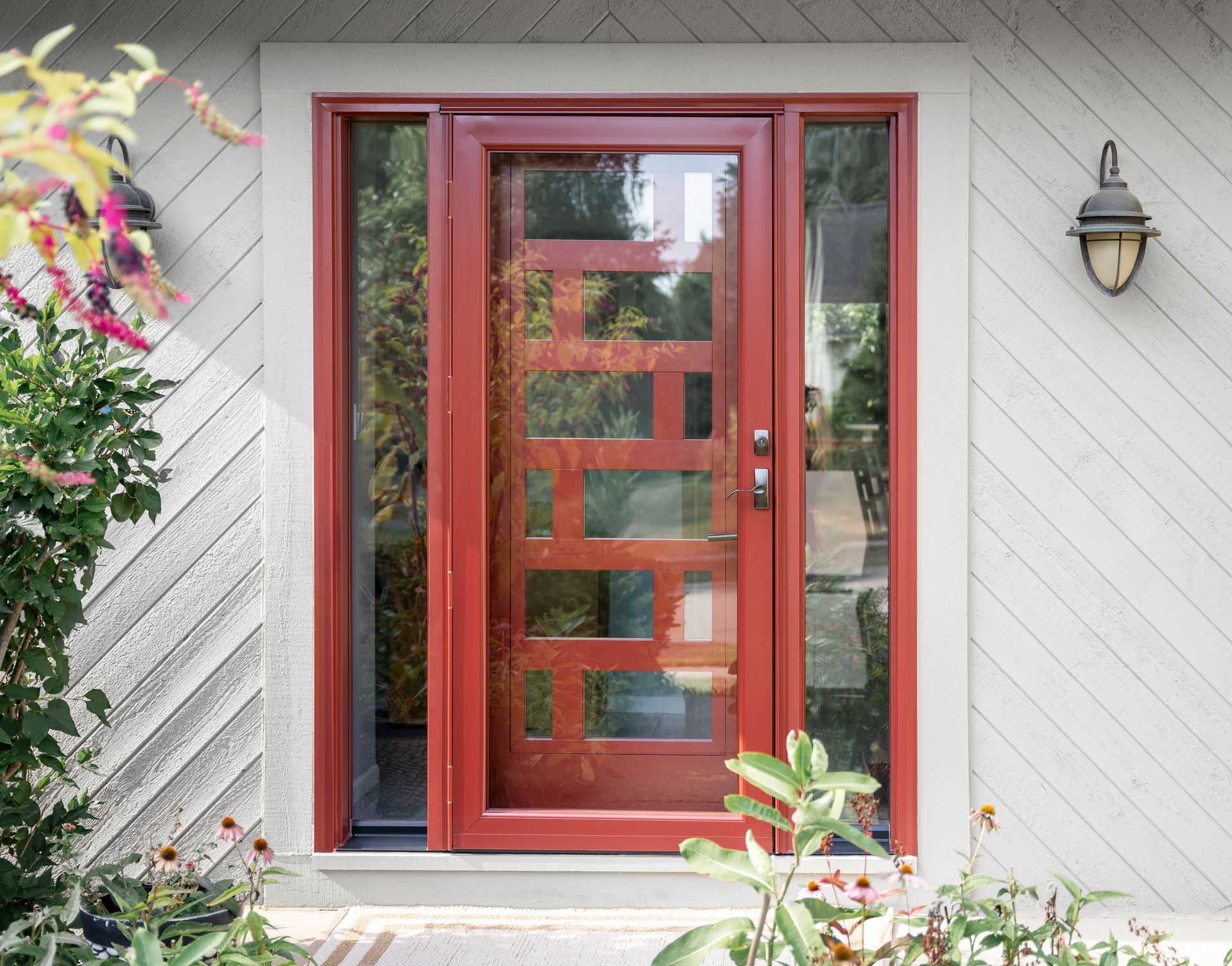 Red front door with glass panels in a white siding exterior, framed by plants and a wall lantern.