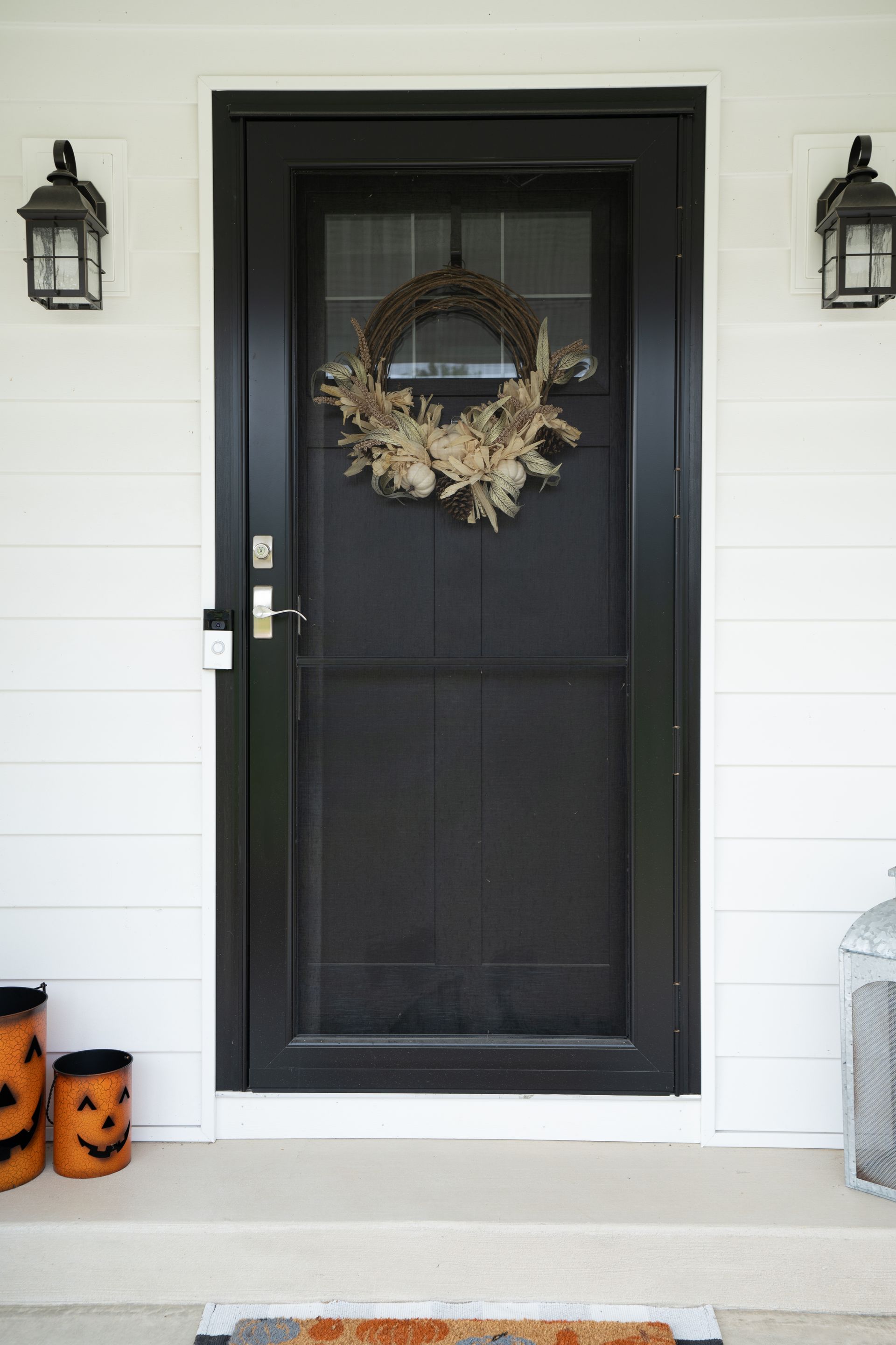 Black front door with fall wreath, flanked by lanterns and Halloween pumpkin decorations on a porch.