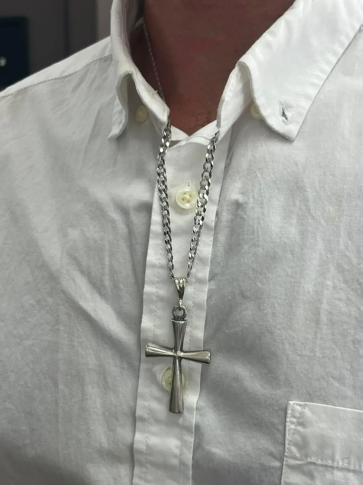 A close-up of a silver cross pendant on a chain, resting against a crisp white button-down shirt.