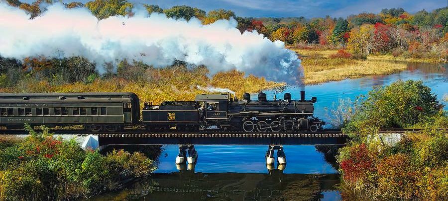 A steam train crossing a bridge over water with trees with fall foliage surrounding it.