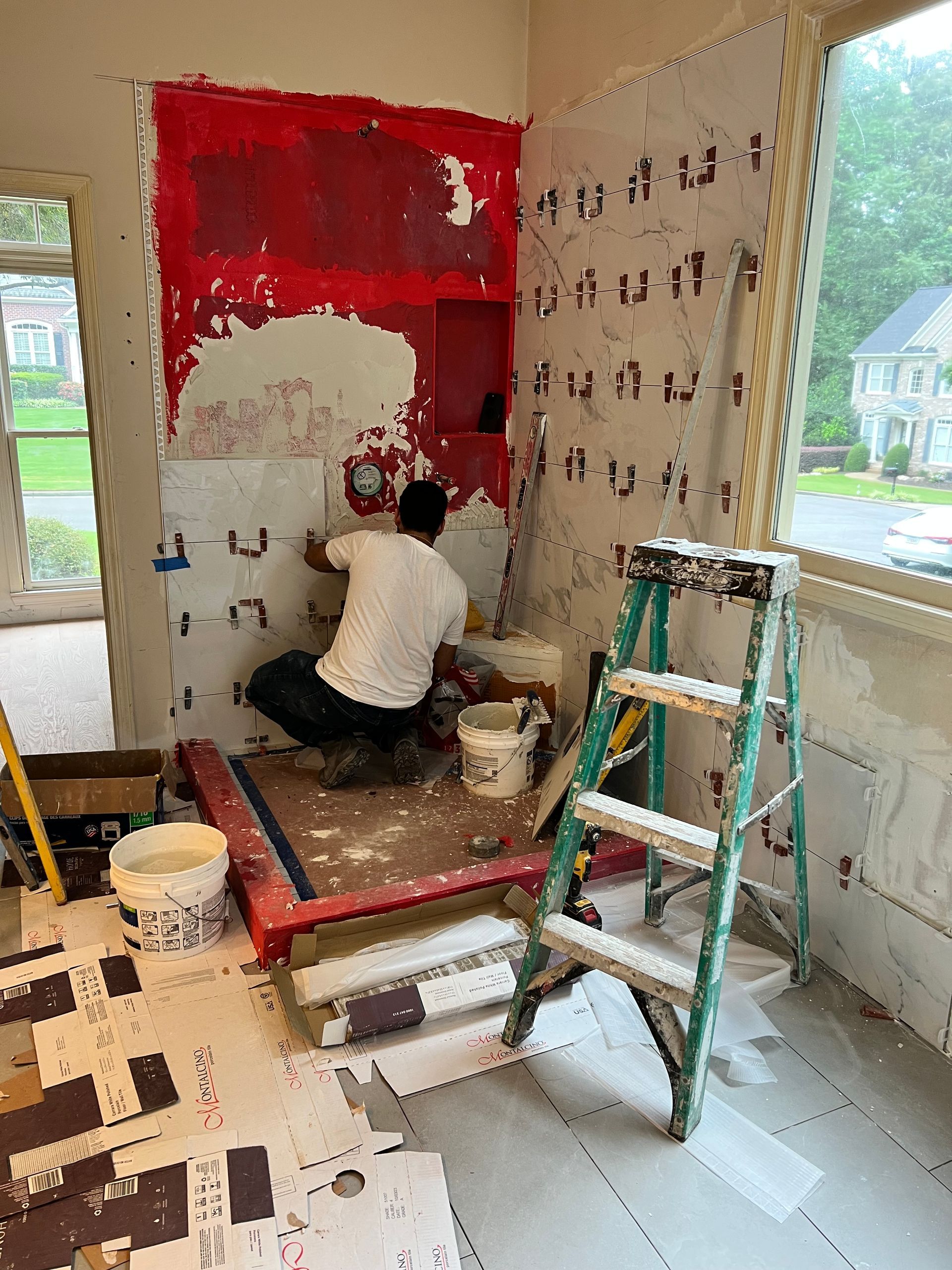 A person installing tile in a bathroom under construction, red wall, green ladder, and white bucket.