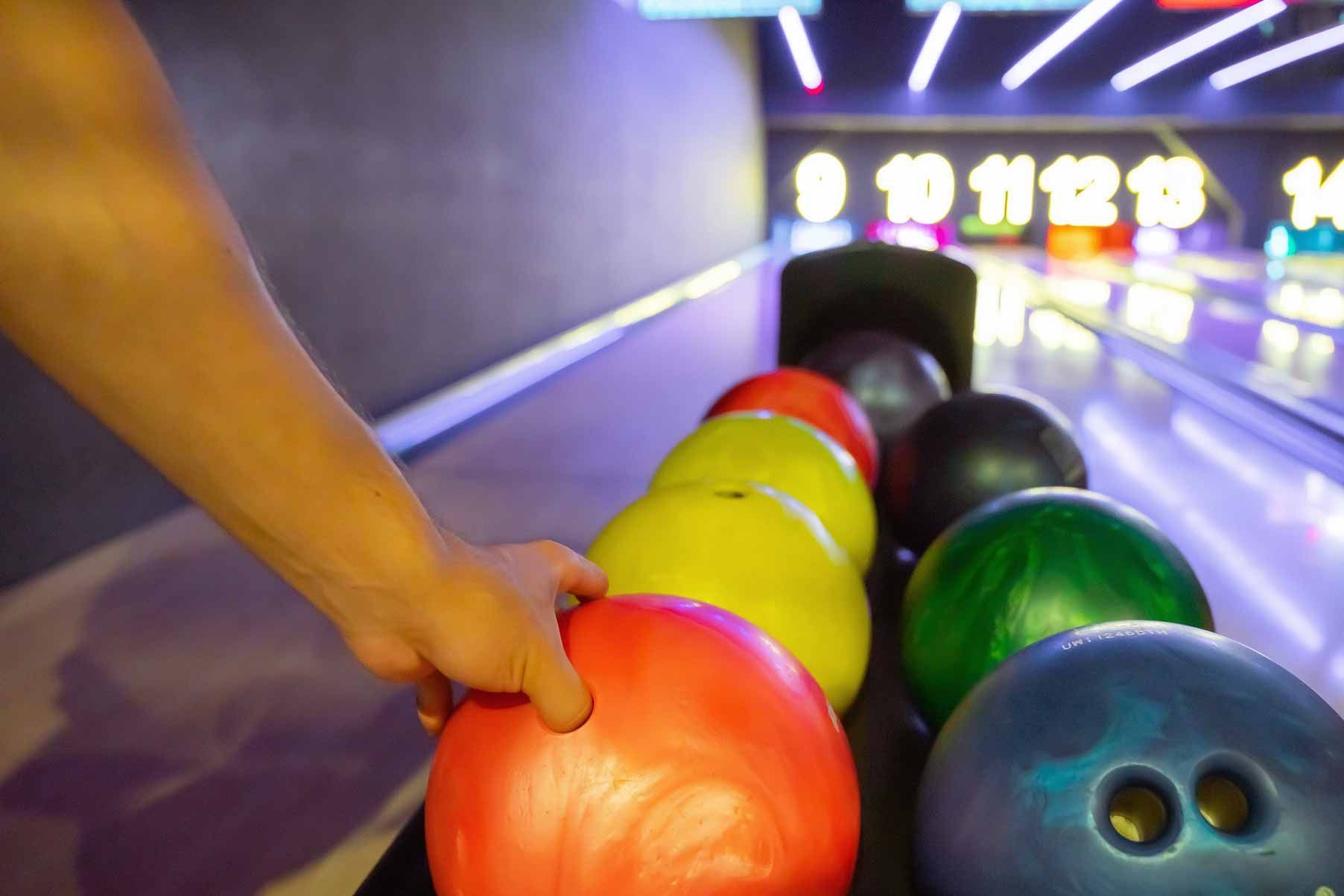 Hand placing a red bowling ball on a bowling lane beside green and yellow balls