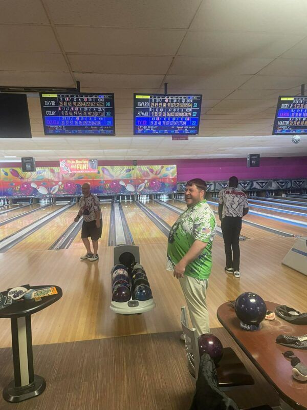 Bowling alley with players near the lanes, scoreboards overhead, and balls on racks in the foreground.