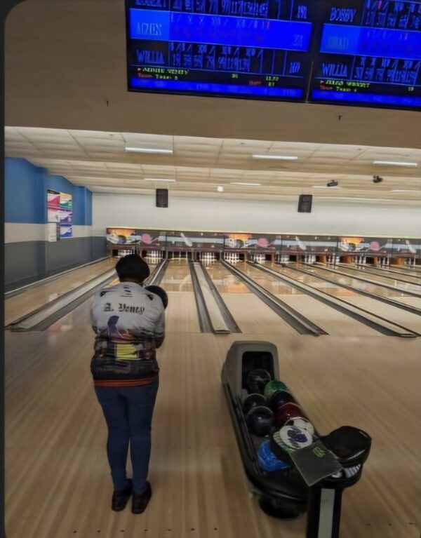 Bowling alley with a person standing at the lane, scoreboards overhead, and bowling balls on the rack.