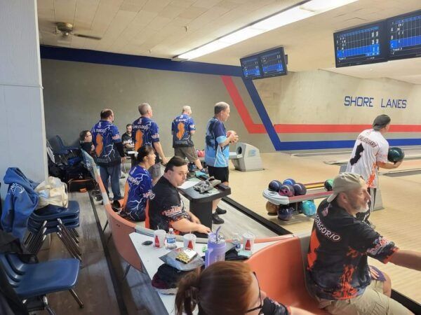 Bowling alley seating area with several people in blue jerseys and others waiting near the lanes