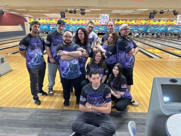 Bowling team posing in a lane, wearing matching purple shirts and holding a peace sign.