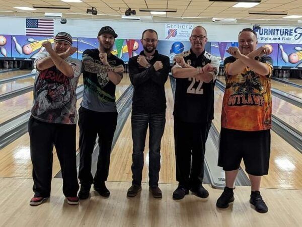 Five men posing at a bowling alley, crossing their arms and smiling in front of the lanes.