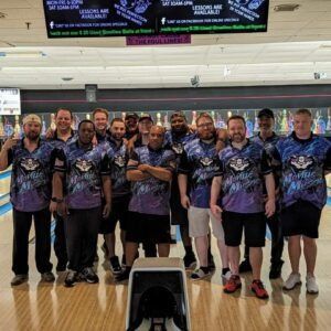 Group of bowling team members posing on a lane, wearing matching purple-and-black shirts.