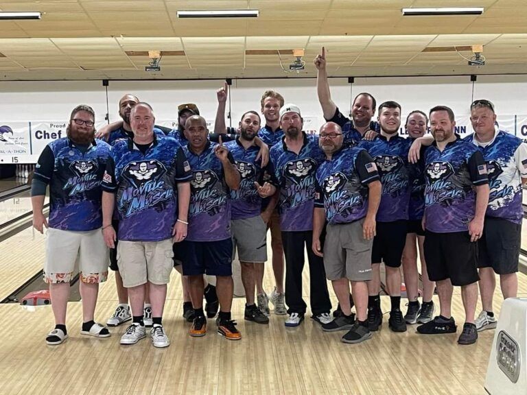Bowling team posing on a lane, wearing matching blue and purple jerseys and smiling with raised hands