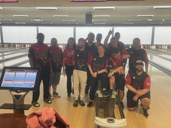 Bowling team posing at the lanes, with a score monitor in front and bowling shoes on the floor.