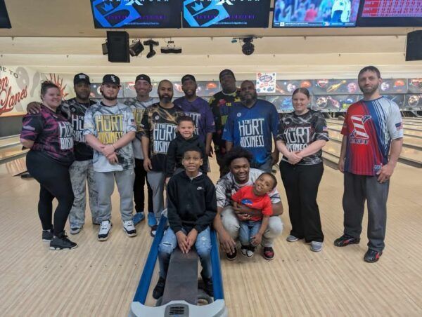 Group photo in a bowling alley, with people posing around a seated boy in the foreground.