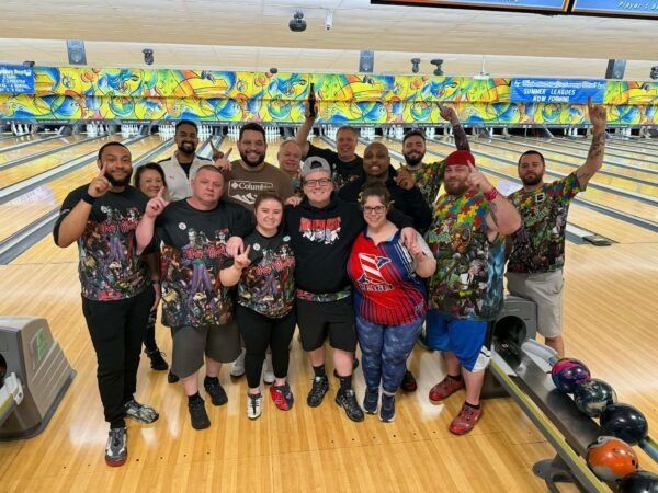 Group of people posing at a bowling alley, smiling with bowling balls and pins in the background.