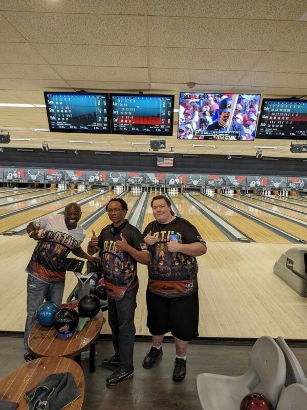 Three bowlers posing at a bowling alley with lanes and scoreboards in the background.