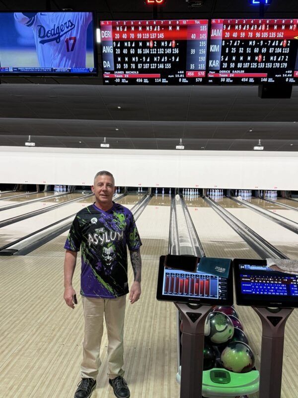 Man standing on a bowling lane with scoreboards and a bowling ball return behind him