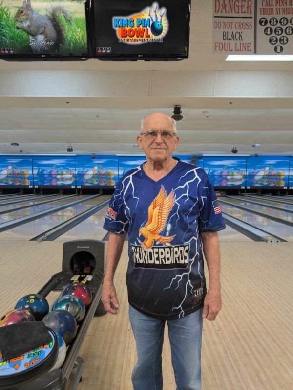 Man standing in a bowling alley holding a bowling ball, wearing a blue Thunderbirds jersey.