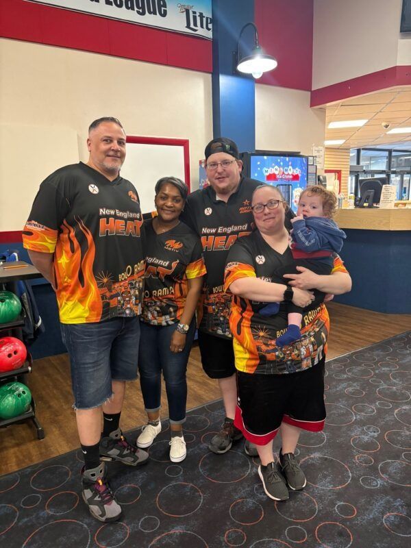 Four people in matching black-orange shirts posing at a bowling alley, with a bowling lane and arcade behind them.