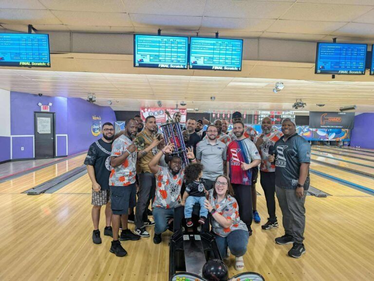 Group of people posing with bowling balls in a bowling alley, with lanes and score screens behind them.