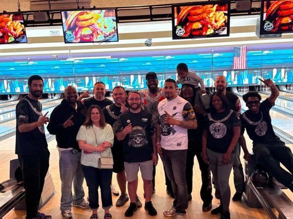 Group of people posing together at a bowling alley, smiling on the lanes with score screens above.
