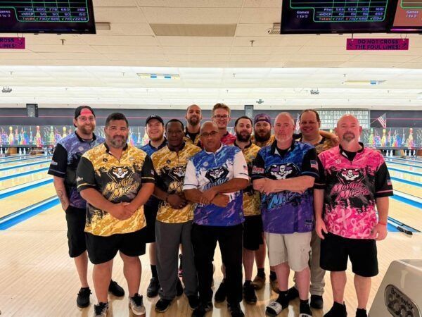 Group of bowlers posing at a bowling alley, wearing colorful jerseys with lanes behind them.