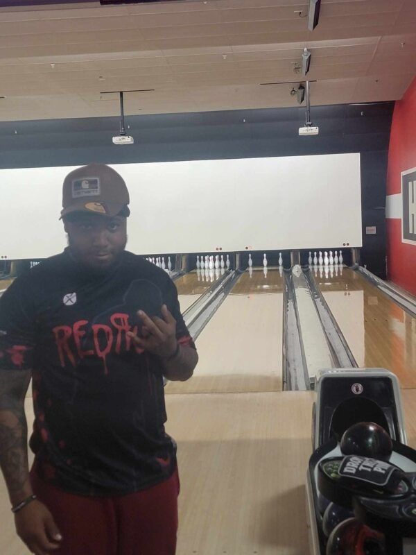 Person standing at a bowling alley, wearing a black Red Sox shirt and cap, with bowling balls in the foreground