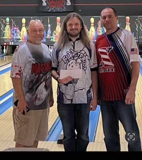 Three men posing in a bowling alley, with lanes and pins visible in the background.