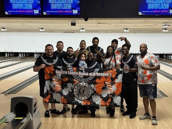 Bowling team posing on lanes, holding a black-and-orange banner in a bowling alley