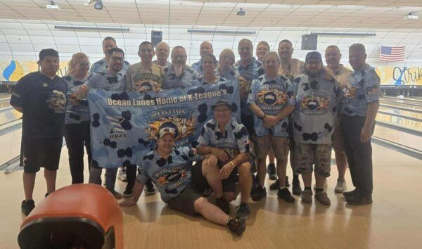 Bowling team posing in a lane, holding a banner and trophies, with pins visible in the background.