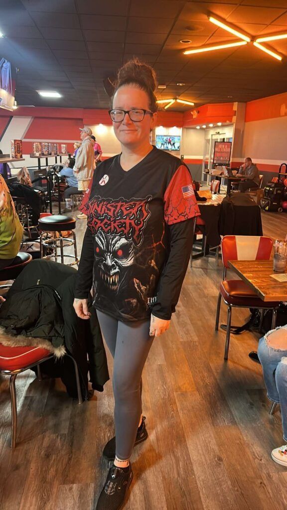 Person standing in a casual restaurant, wearing a black graphic shirt and gray pants under orange ceiling lights.