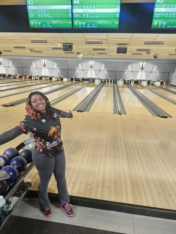 Bowling alley with a smiling person posing beside a ball return and lanes behind them