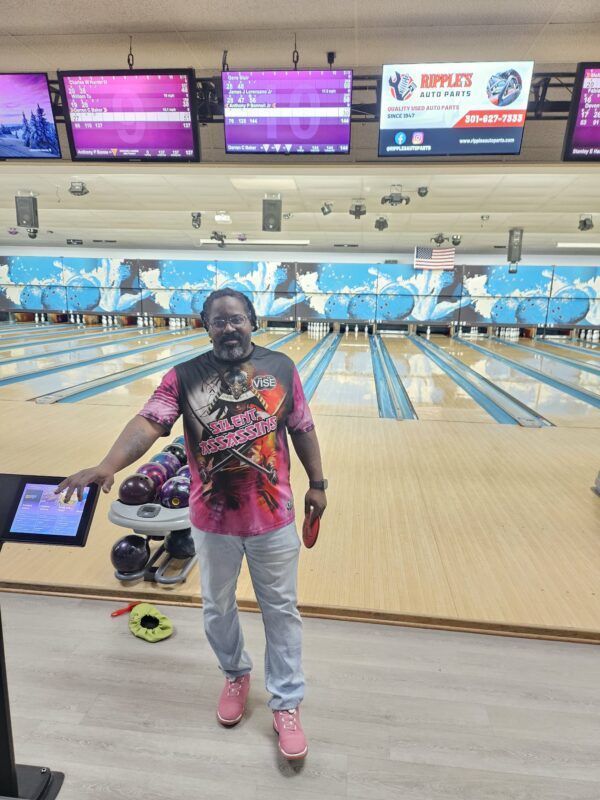 Person posing at a bowling alley, holding a bowling ball bag, with lanes and scoreboards behind.