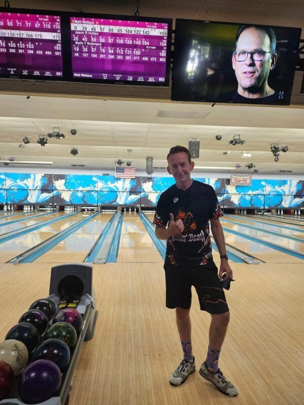 Bowling alley with a man holding a bowling ball, lanes, ball rack, and score screens in the background