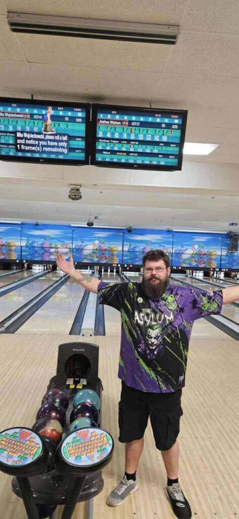 Man posing with arms out at a bowling alley, lanes and scoreboards behind him.