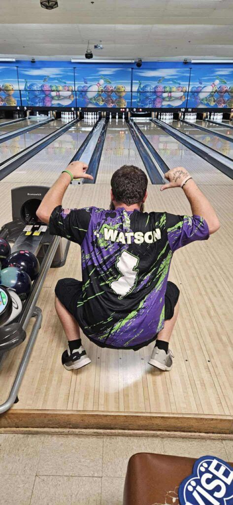 Bowler crouching at the lane approach in a purple WATSCO shirt, with bowling lanes and pins ahead