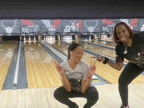 Two women posing and smiling on a bowling lane, with bowling pins in the background.