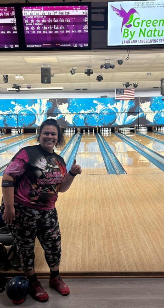 Person posing in a bowling alley with blue lanes, pins, and a Green by Nature sign in the background