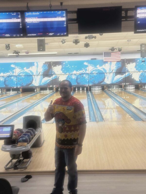Person standing in a bowling alley, holding a drink and giving a thumbs-up near the lanes.