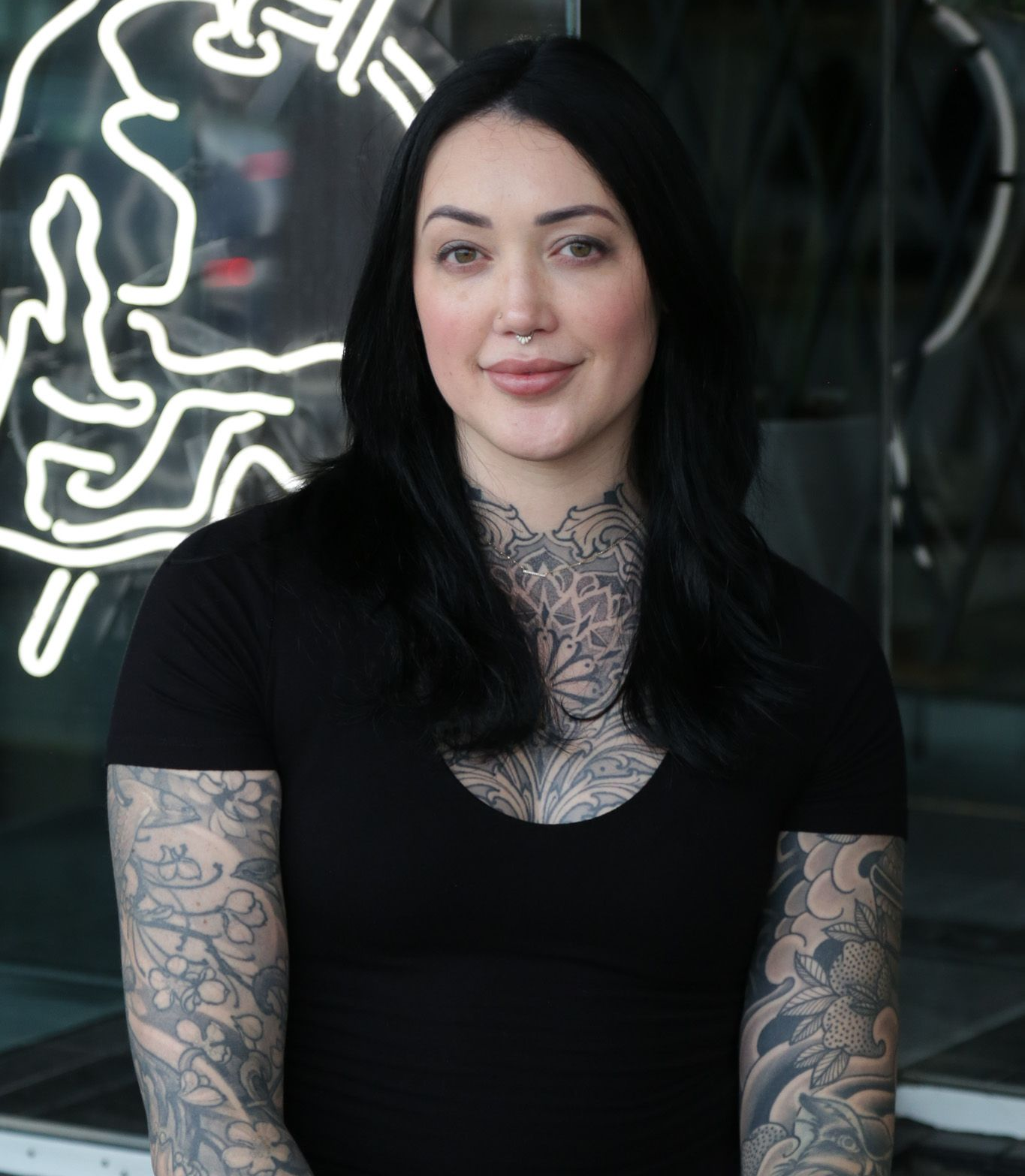 Woman with tattoos on arms and chest stands in front of a neon sign.