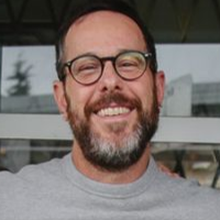 Smiling man with glasses and a beard, wearing a gray shirt, standing outdoors near a building.