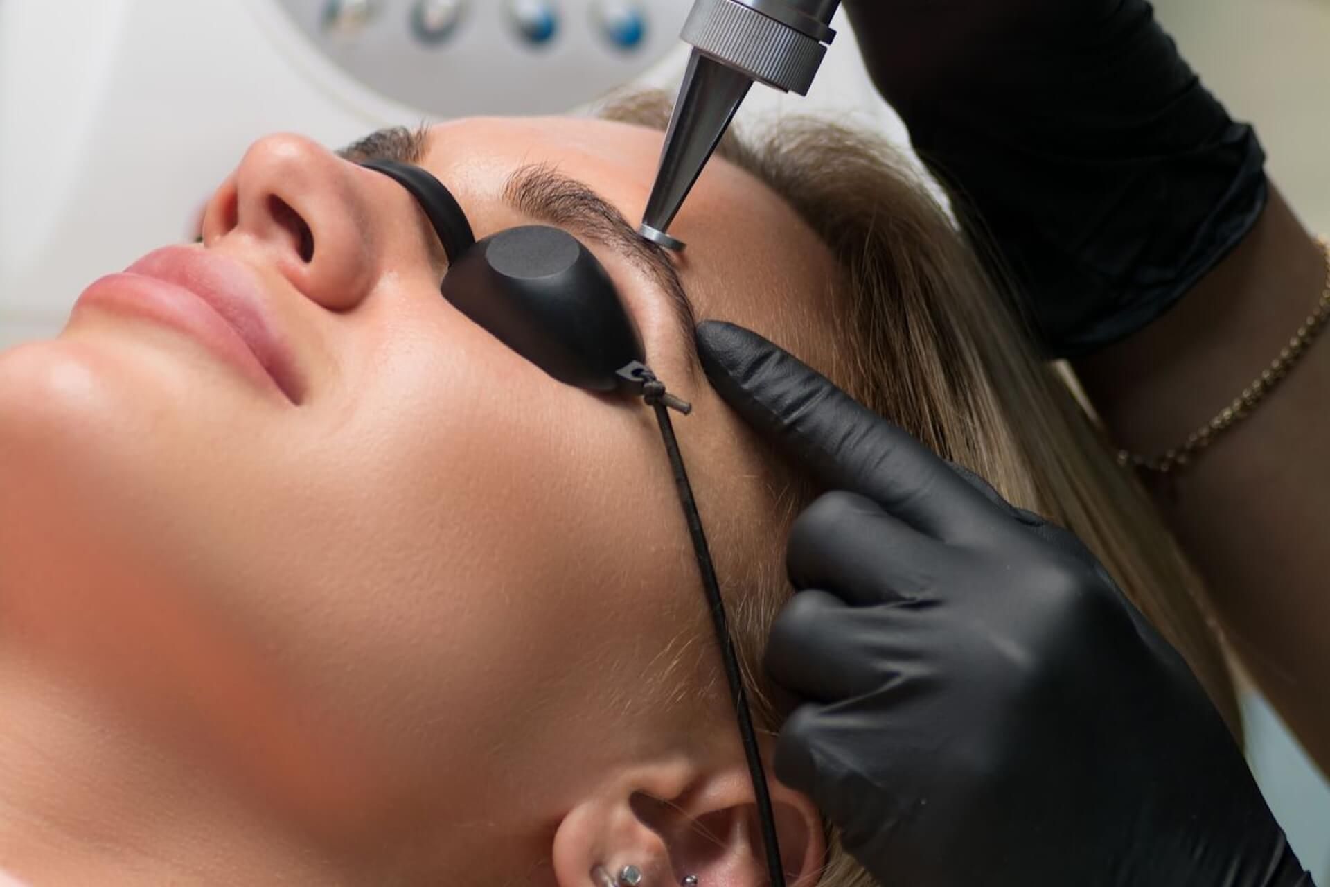 Cosmetic procedure on a client’s eyebrow with a handheld tool and gloved hands