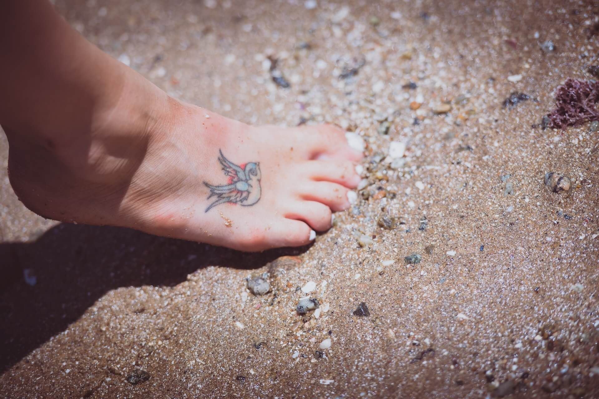 Bare foot with a small tattoo resting on sandy, pebbled ground in sunlight