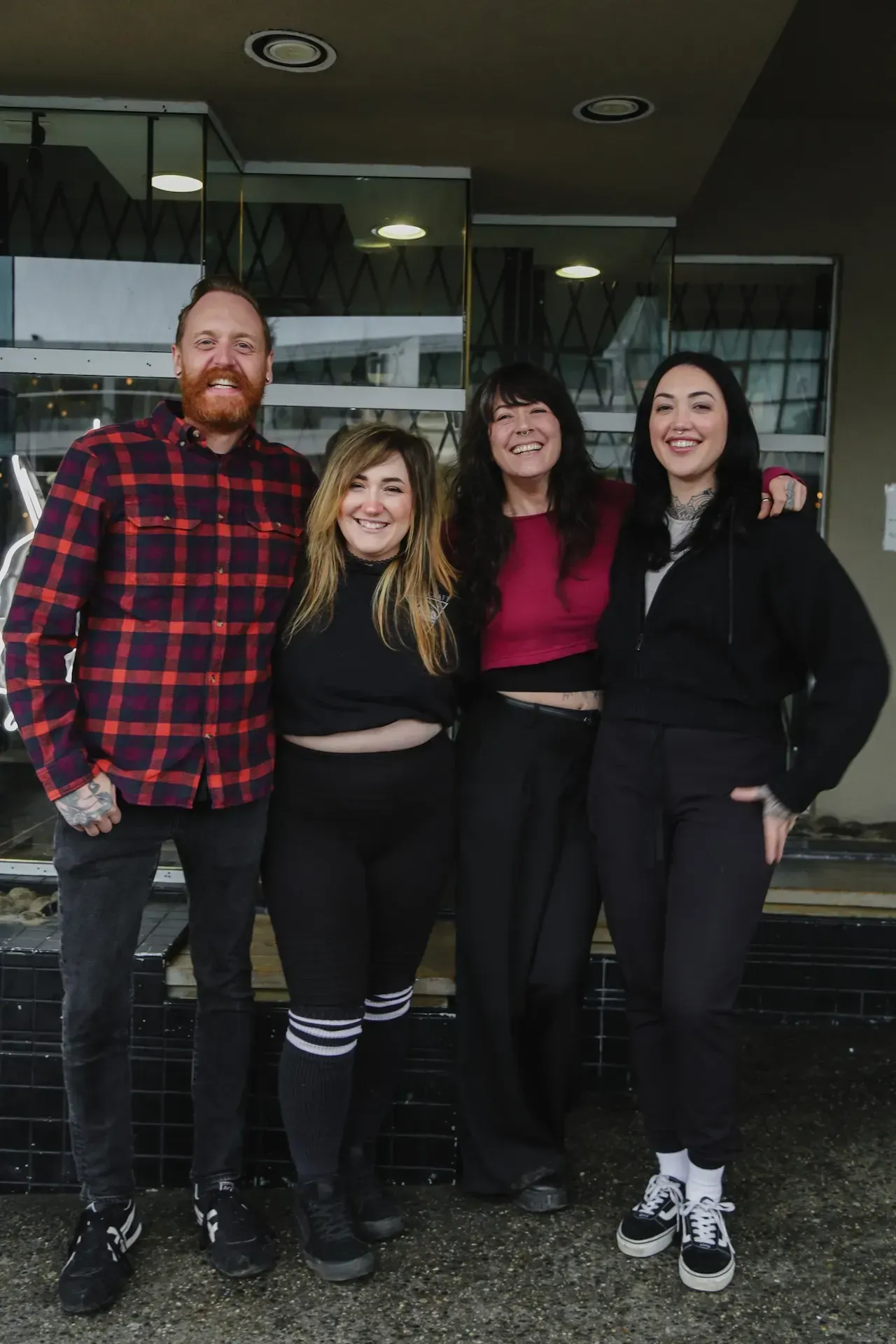Four smiling people stand side-by-side outdoors in front of a building with glass doors and reflective windows.
