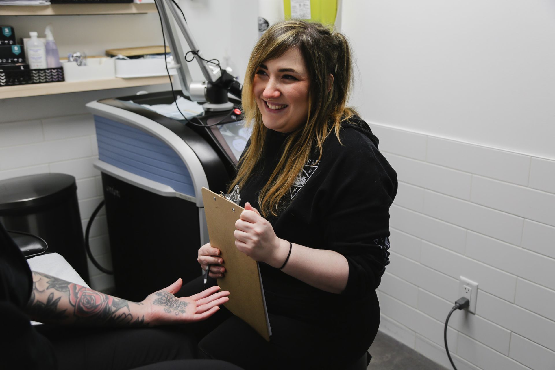 Smiling woman holding a clipboard in a tattoo studio beside a seated client with a tattooed arm