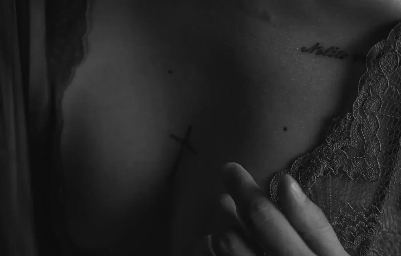 Close-up of a person's chest, with a cross tattoo and a small word tattoo.  Hand touches the chest.