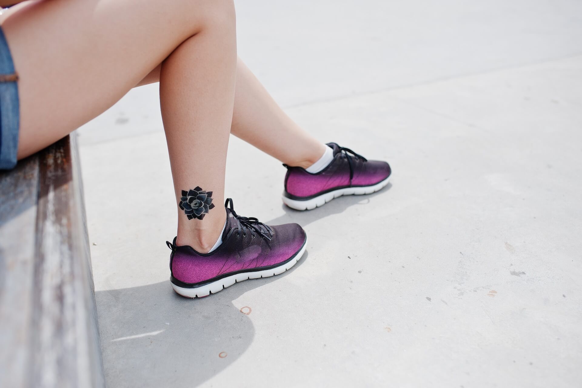 Legs resting on a bench, wearing purple running shoes with black laces and a floral ankle tattoo.