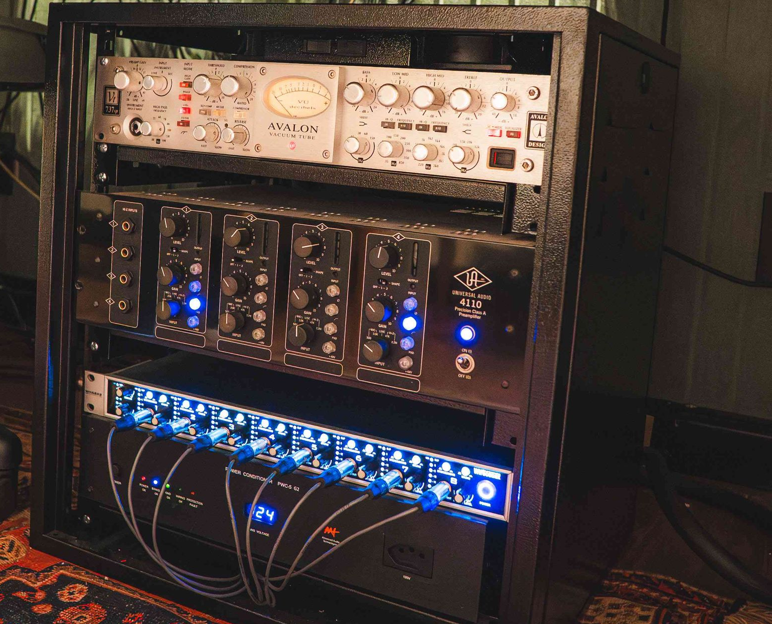 Um rack de equipamentos preto está sobre um tapete estampado embaixo de uma mesa, contendo várias unidades de áudio de estúdio com luzes acesas.