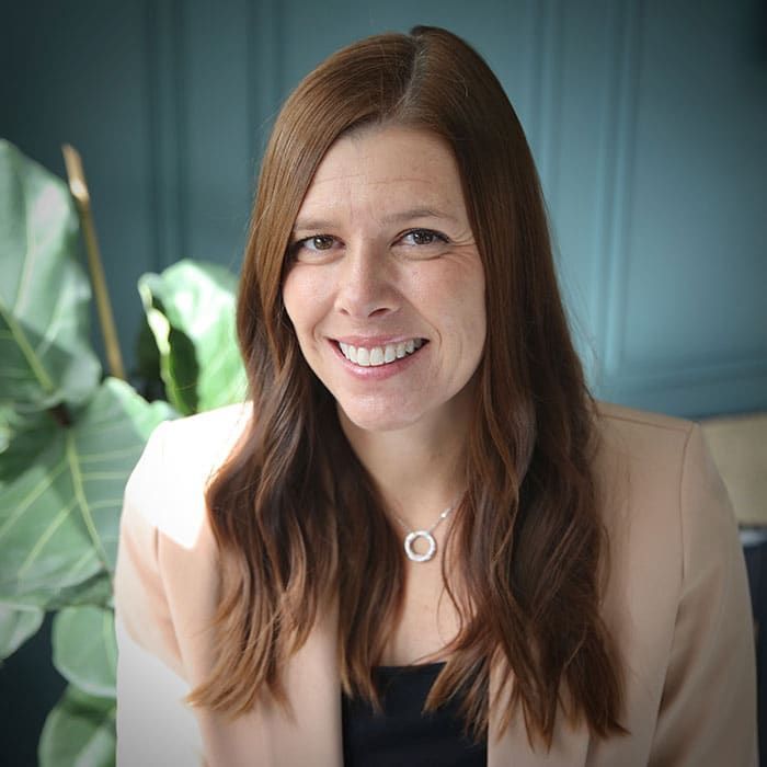 Woman with brown hair smiles, wearing a cream blazer and a necklace. Background is a green plant and a teal wall.